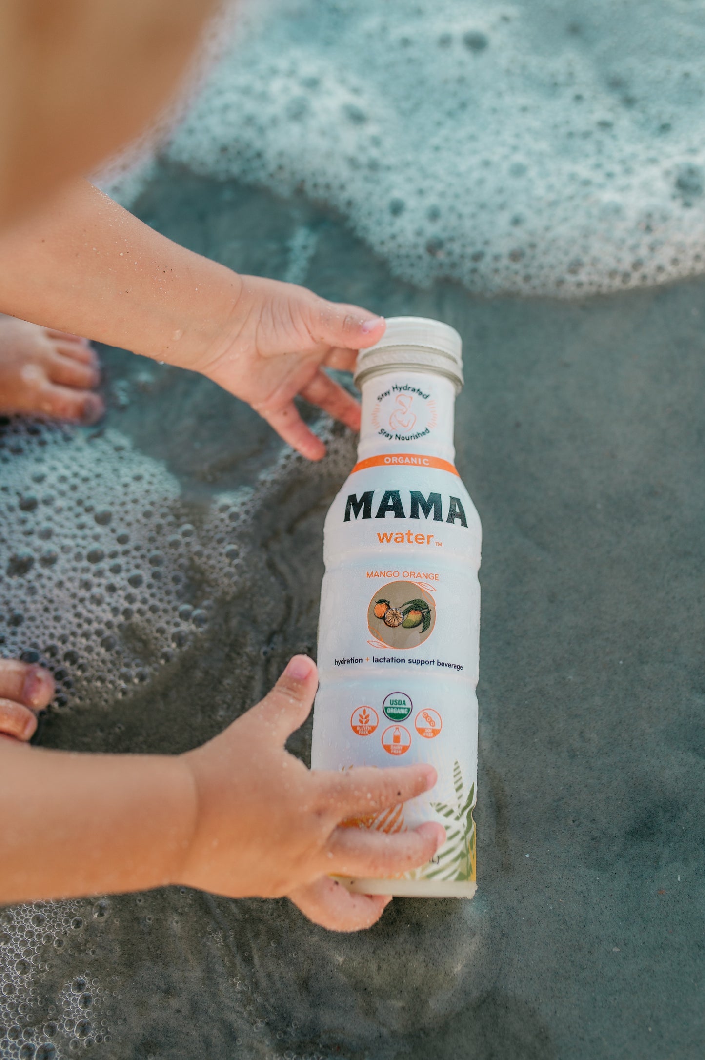 Toddler holding a 'MAMA water' bottle by a body of water with bubbles.