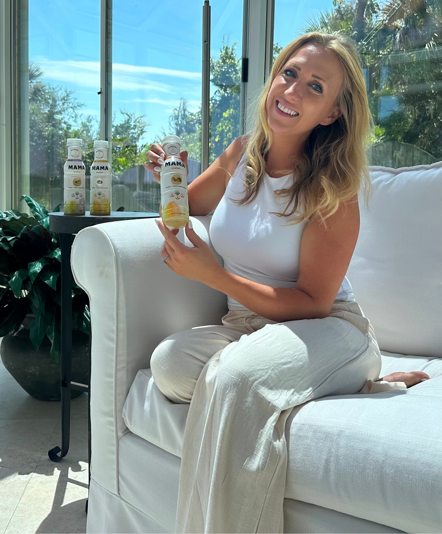Amanda sitting on a white couch holding a bottle of a product, with more bottles on a surface in the background.