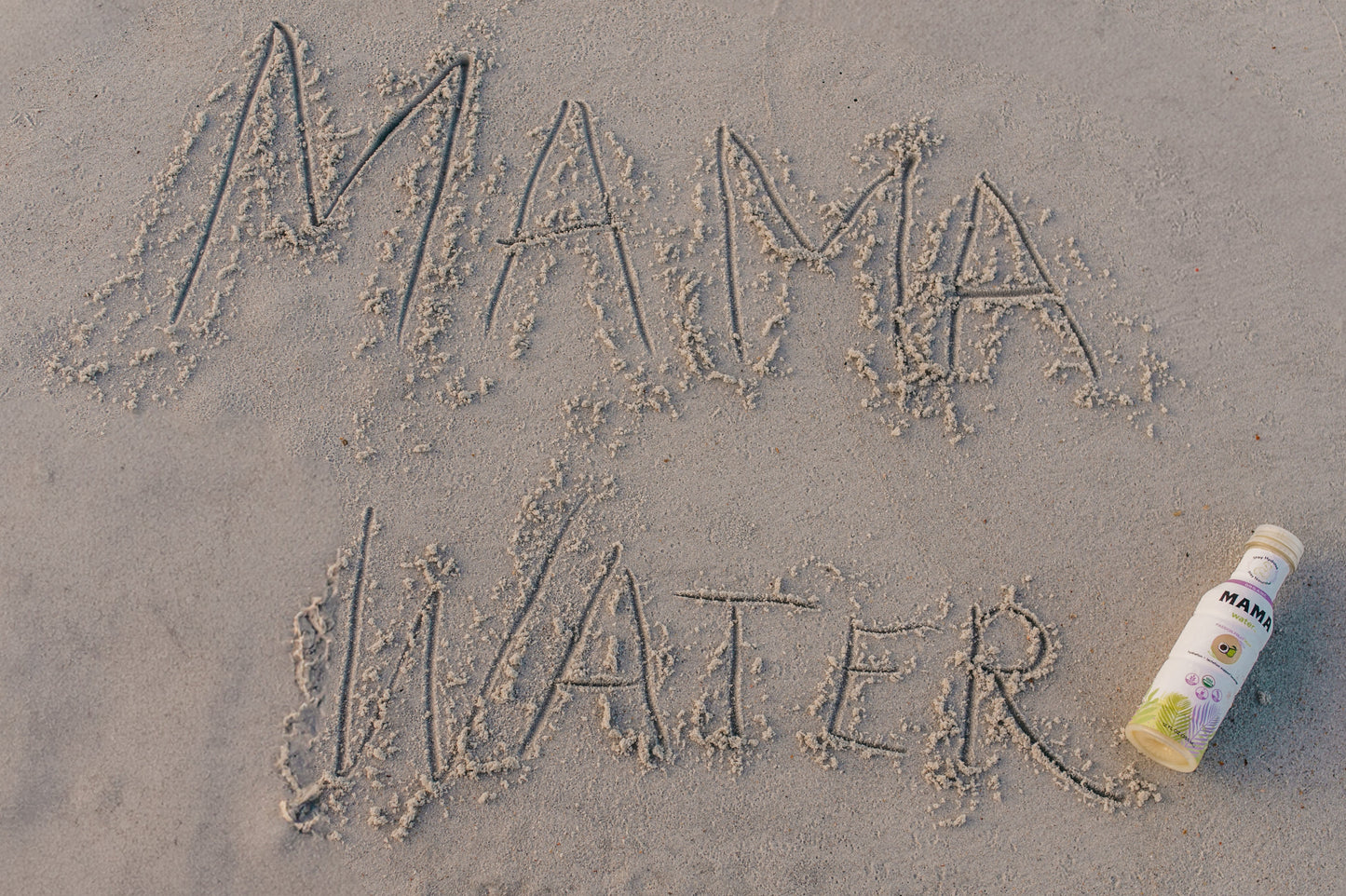 Bottle labeled 'Mama Water' on sand with 'MAMA WATER' written in sand.